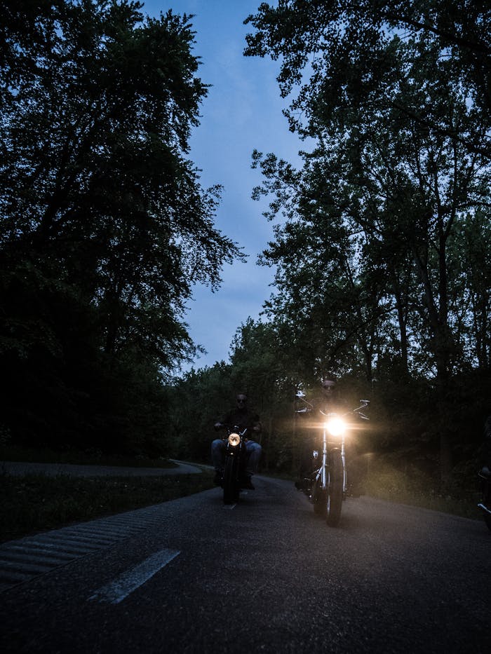 Two motorcyclists ride through a tree-lined road at dusk in Soest, Netherlands, with headlights on.